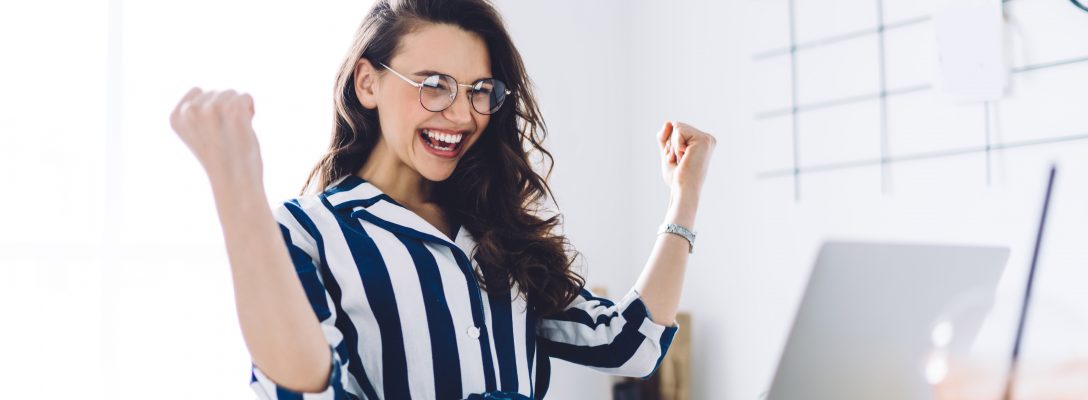 Happy young woman sitting at table with hands up in winner gesture Locking down a new role during COVID